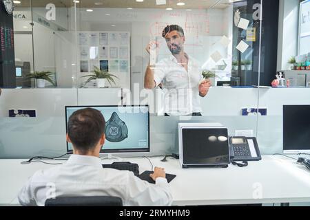 Rückansicht eines anonymen männlichen Arbeiters, der eine Skizze der covid-Maske vorbereitet, während der Kollege ein Schema auf Glaswand im Labor zeichnet Stockfoto