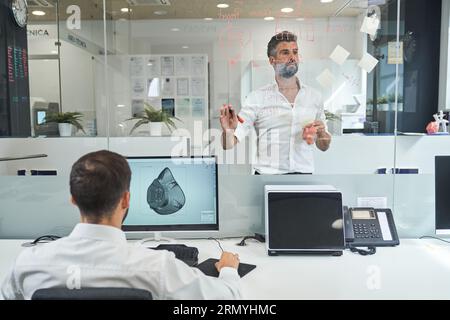 Rückansicht eines anonymen Mannes, der Computer benutzt und ein grafisches Maskenmodell aus der Coronavirus-Pandemie herstellt, während er im Büro mit männlichen Kollegen arbeitet Stockfoto