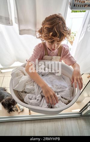 Von oben ein fokussiertes Kind, das weiße Laken an der Wäscheleine aufhängt, während es zu Hause Haushaltsroutinen auf dem Balkon macht Stockfoto