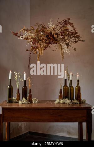 Altmodisches Layout, bestehend aus Glasflaschen mit beleuchteten Kerzen und Ästen innen und weißen Blumen auf braunem Holztisch unter hängendem b Stockfoto