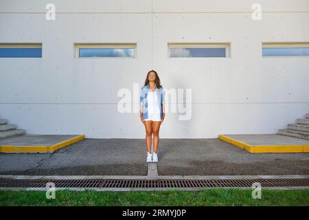 Ein ganzer Körper einer jungen Frau in lässigem Outfit, die vor dem modernen Gebäude steht, mit geschlossenen Augen Stockfoto