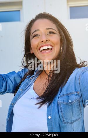 Lachende junge Frau mit langen Haaren in lässiger Kleidung und Blick weg auf sonnige Straße Stockfoto
