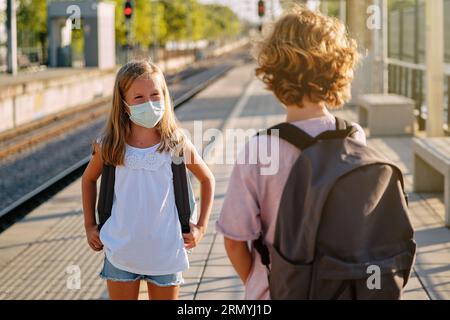 Junge und Mädchen in Schutzmasken, die einander anschauen und sprechen, während sie auf den Zug auf dem Bahnsteig während der Coronavirus-Epidemie warten Stockfoto