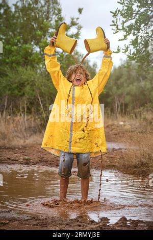 Ein Kind in voller Länge im gelben Regenmantel schreit und gießt schmutziges Wasser aus Gummistiefeln, während es an regnerischen Tagen in einer Pfütze im Wald steht Stockfoto