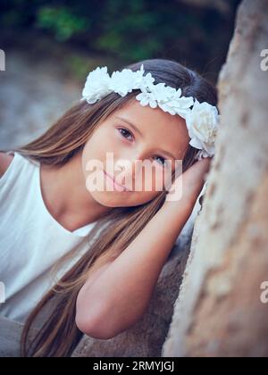 Friedliches, süßes, junges Kind mit weißem Blumenkranz und elegantem Kleid, das sich an die Wand lehnt, während es in die Kamera schaut Stockfoto