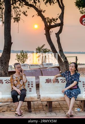 Pakse, Süd-Laos-Februar 12 2023: Sitzen auf chinesischen beschrifteten Metallbänken, in der Nähe des Flusses, die Damen entspannen und geselligen sich, während die Dämmerung naht, en Stockfoto
