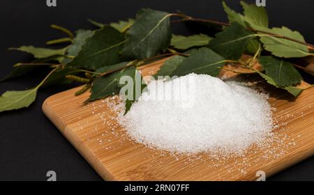 Nützliches Süßungsmittel Xylit, Birkenzucker auf Holzbrettern, Stäbchen, Birkenblätter, dunkler Hintergrund. Gesunde Ernährung. Flach verlegt. Horizontal Stockfoto