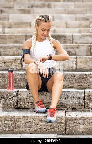 Läuferin überprüft die Ergebnisse von Smartwatches, sitzt auf der Treppe, nimmt nach dem Joggen und Ruhen den Puls, Armband mit Mobiltelefon. Stockfoto