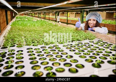 Fröhliche Gärtnerin, die ihre Zustimmung zeigt und lächelt, während sie sich auf das Regal lehnt und Pflanzen im Gewächshaus sät. Frau mit Gartenhandschuhen und Einwegmütze. Stockfoto