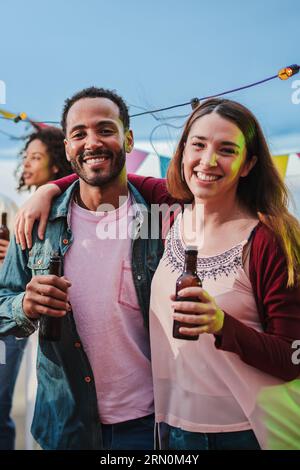 Vertikales Porträt eines glücklichen Paares, das lächelt und die Kamera auf einer Nachmittagsfeier mit ihren multirassischen Freunden anschaut. Ein Paar Freunde, die Bierflaschen halten und gemeinsam auf einem lustigen fest trinken. Hochwertige Fotos Stockfoto