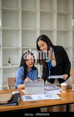 Zwei professionelle und schöne asiatische Geschäftsfrauen oder Buchhalterinnen diskutieren über die Arbeit und prüfen zusammengefasste Finanzdaten auf einem Tablet Stockfoto