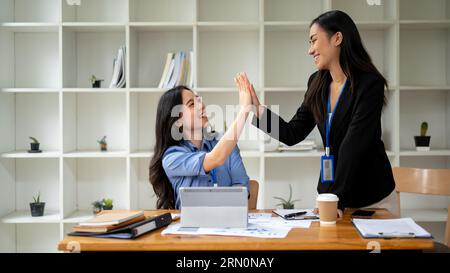 Zwei schöne und fröhliche asiatische Geschäftsfrauen geben sich gegenseitig High Fives, während sie gemeinsam an einem Projekt im Büro arbeiten. Aufmuntern, Teamwork Stockfoto