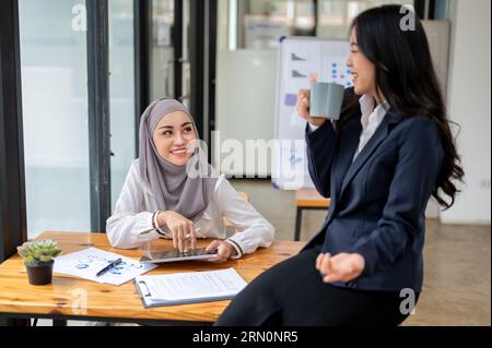 Eine attraktive und lächelnde asiatisch-muslimische Büroangestellte, die einen Hijab trägt, unterhält sich gerne während einer Kaffeepause im Büro mit ihrer Kollegin. Stockfoto