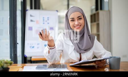 Ein Porträt einer schönen, lächelnden asiatischen muslimischen Geschäftsfrau, die einen Hijab trägt, sitzt an ihrem Schreibtisch in einem modernen Büro. Stockfoto