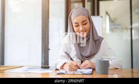 Eine attraktive asiatische muslimische Geschäftsfrau, die einen Hijab oder Kopftuch trägt, füllt Formulare aus, unterzeichnet Dokumente oder macht Notizen auf Papier an ihrem Schreibtisch Stockfoto