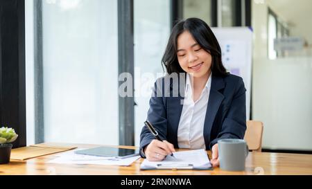 Eine attraktive asiatische junge Geschäftsfrau füllt Formulare aus, unterzeichnet Dokumente oder macht Notizen auf Papier an ihrem Schreibtisch in einem modernen Büro. Stockfoto