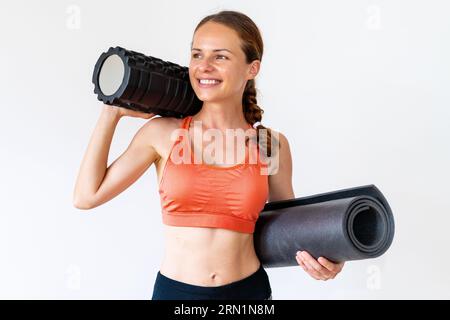 Junge Athletin schlanke Frau in Sportbekleidung, die Schaumstoffrolle und gerollte Yogamatte in den Händen hält und lächelt, Stockfoto