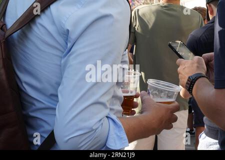 Junger Mann mit zwei Plastikgläsern mit Bier in jeder Hand, der während des Sommerfestes unter anderen steht. Stockfoto