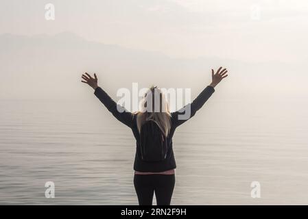 Reisende Frau mit erhobenen Armen, die an einem nebeligen Tag vor einem See steht. Stockfoto