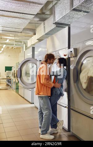 Seitenansicht eines jungen, romantischen, multiethnischen Paares, das sich in der Nähe von Waschmaschinen in der öffentlichen Wäscherei umarmt Stockfoto