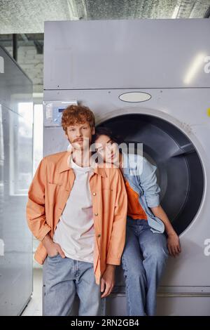 Junge asiatische Frau, die auf der Waschmaschine in der Nähe eines Freundes in der öffentlichen Wäscherei sitzt Stockfoto