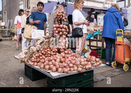 Ein Stapel frischer Zwiebeln, während die Kunden sich anstellen, um an einem Stand auf dem Wochenmarkt in Plougasnou, Britanny, Frankreich, festliches Gemüse zu kaufen. Stockfoto