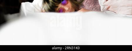 Eine Frau, die eine Sonnenbrille trägt, entspannt sich auf der zerknitterten weißen Bettwäsche in sanften Lichtern, komfortablem Schlafzimmer, Entspannungszeit, Hotel- oder Bettenbusi Stockfoto
