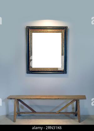 Ein leerer Leinwandbilderrahmen, der an der Wand hängt, mit Clipping-Pfad, eine Holzbank gegen die blau-graue Farbwand, gepunktete Beleuchtung von oben Stockfoto
