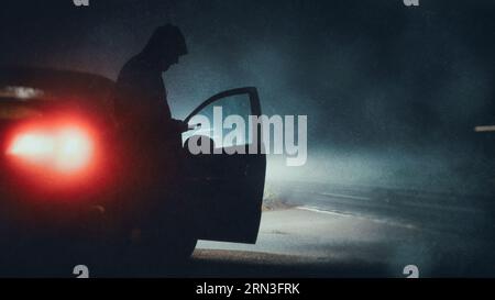 Eine lustige, geheimnisvolle Figur mit Kapuze, die nachts neben einem Auto steht. Ein Telefon. Scheinwerfersilhouette. Mit einem grunge, abstrakten unscharfen Schnitt Stockfoto