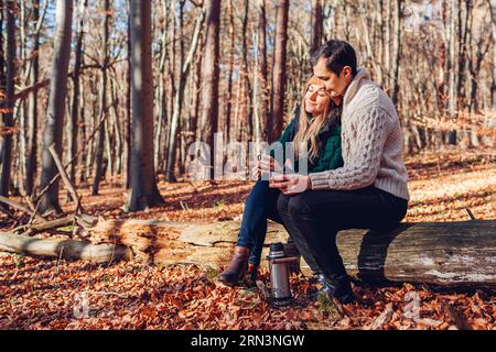 Porträt eines jungen Liebespaares, das Tee trinkt und sich im Herbstwald entspannt. Mann und Frau sitzen auf dem Kofferraum und genießen die herbstliche Landschaft Stockfoto