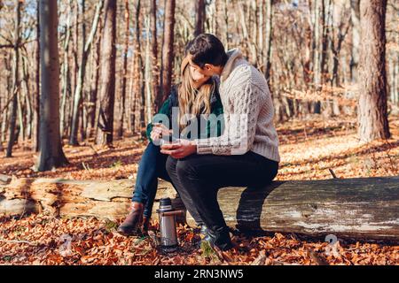 Glückliches Paar, das sich küsst, Tee aus Thermoskannen trinkt und sich im Herbstwald entspannt. Mann und Frau sitzen auf dem Kofferraum und trinken Kaffee aus dem Vakuumbehälter. Menschen Stockfoto