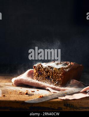 Geschnittenes Bananenbrot auf einem rustikalen Holztisch und schwarzem Hintergrund. Etwas Zuckerpulver in der Luft, das im Bild Bewegung erzeugt. Vertikales Stillleben Stockfoto