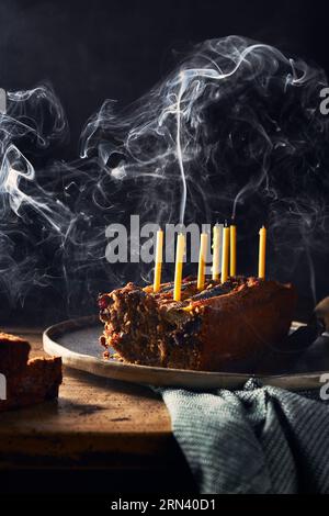 Geschnittenes Bananenbrot auf einem rustikalen Holztisch und schwarzem Hintergrund. Es hat Geburtstagskerzen darin, die noch rauchen. Serviette auf dem Tisch. Vertikal Stockfoto