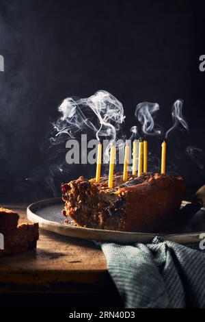 Geschnittenes Bananenbrot auf einem rustikalen Holztisch und schwarzem Hintergrund. Es hat Geburtstagskerzen darin, die noch rauchen. Serviette auf dem Tisch. Vertikal Stockfoto