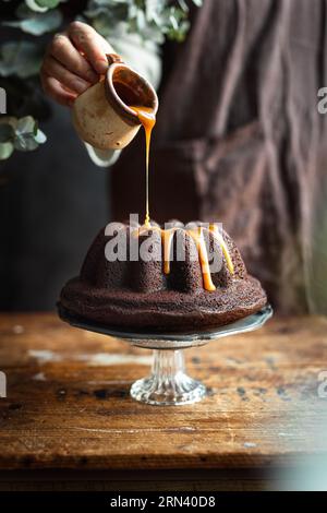 Person im Hintergrund, die Karamell über Schokoladenkuchen gießt. Rustikale Einrichtung. Der Kuchen steht auf dem eleganten Glastisch auf einem alten Holztisch. Stockfoto
