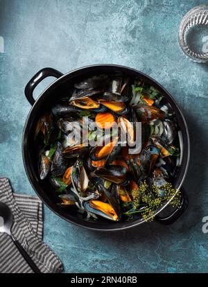 Flachbild von oben von gedämpften und verzehrfertigen Muscheln im schwarzen Topf. Blauer strukturierter Hintergrund. Glas Wasser, Löffel und Serviette A Requisiten. Stockfoto