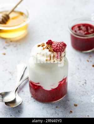 Nahaufnahme von Joghurt-Parfait mit Granola und Himbeermarmelade. Weißer Steinhintergrund mit Krumen aus Granola, Löffeln, Honig im Glas und Himbeermarmelade Stockfoto