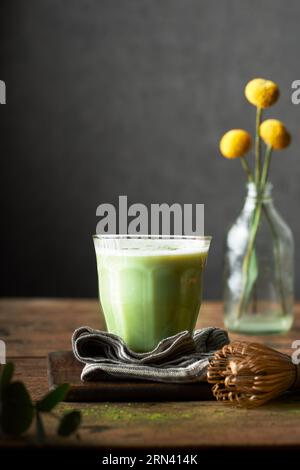 Ein Glas Matcha Latte auf einem rustikalen Holztisch und grauem Hintergrund. Einrichtung im Innenbereich. Es gibt Blumen, Serviette, Schneidebrett und Bambusbesen. Stockfoto