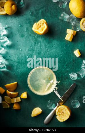 Zitruscocktail auf blauem Hintergrund, umgeben von Eis, Spritzern und Zitronen. Einige Zitronen sind bereits eingedrückt. Zubereitung und Zutaten. Stockfoto