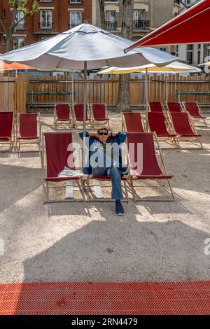 Paris, Frankreich - 08 27 2023: Strände Von Paris. Eine reife Frau, die auf einem Liegestuhl sitzt, unter einem weißen Sonnenschirm an den Kais des Bassin de la Villette Stockfoto