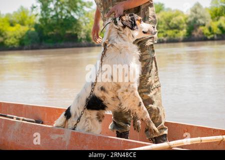 Fischer in Tarnkleidung, der nach dem Angeln nassen Hund aus dem Holzboot zieht. Stockfoto
