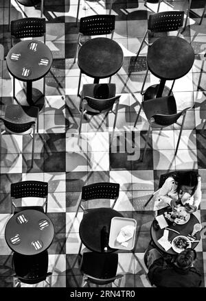 Gäste in der Union Station Food Hall, Washington DC — Schwarz-Weiß-Foto | WASHINGTON D.C., Vereinigte Staaten — Gäste teilen sich einen Tisch in der Union Station Food Hall, gesehen in einer Schwarz-Weiß-Aufnahme aus der Vogelperspektive eines Schachbrettfliesenbodens, der mit Stuhlschatten gemustert ist. Die Essenshalle der Union Station befindet sich teilweise in dem 1907 erbauten Beaux-Arts-Terminal, das heute ein beliebter Halt für Reisende und Pendler ist, die täglich durch den Bahnhof fahren. Stockfoto