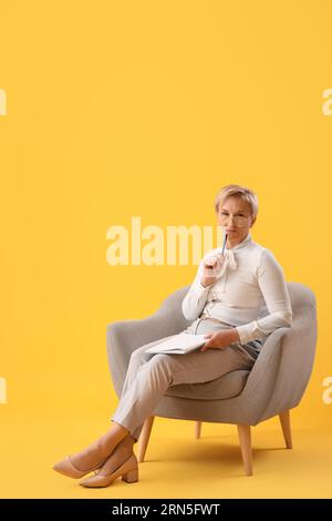Reifer Psychologe mit Notizbuch im Sessel auf gelbem Hintergrund Stockfoto