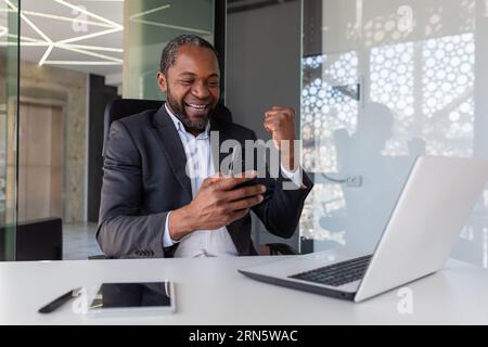 Reife erfolgreiche afroamerikanische Geschäftsmann erhielt online Benachrichtigung über die Gewinnernachricht am Arbeitsplatz im Büro, Mann mit Telefon, mit App auf Smartphone, glücklicher Gewinner mit Erfolg. Stockfoto