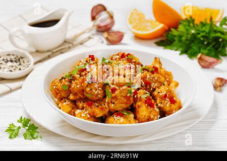 Knusprige Blumenkohlblüten mit Sesamsamen, Chiliflocken und Petersilie in weißer Schüssel auf weißem Holztisch mit Zutaten, horizontal Stockfoto