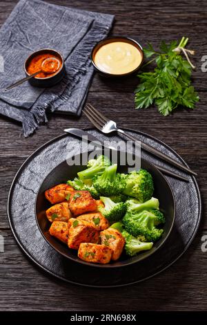 Gebackene Lachsstücke mit gedämpften Brokkoliröschen in schwarzer Schüssel auf dunklem Holztisch mit Sriracha-Mayo-Sauce, vertikale Ansicht von oben Stockfoto