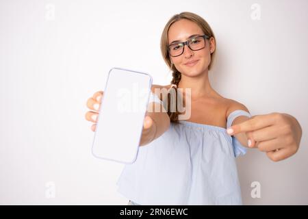 App-Anzeige. Lächelnde hübsche Frau, die das Smartphone mit dem weißen, leeren Gerätebildschirm in der Hand in der Nähe der Kamera hält, oranges Studio. Gadget mit leerem freien Speicherplatz Stockfoto