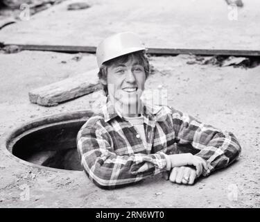 1970ER JAHRE LÄCHELNDER MANN ARBEITER MIT KAROHEMD MIT SCHUTZHELM UND BLICK AUS DEM SCHACHT IN DER STRASSE DER STADT - S20060 HAR001 HARS B&W AUGENKONTAKT FÄHIGKEITEN BERUF GLÜCK FÄHIGKEITEN KOPF UND SCHULTERN KUNDENSERVICE ARBEITSVERHÄLTNISSE BERUFE ZUGANG STÄDTE MITARBEITER LÖSUNGEN JUNGER ERWACHSENER MANN SCHWARZ-WEISS KAUKASISCHE ETHNIE HAR001, DER ALTMODISCHE ÖFFENTLICHE VERSORGUNGSEINRICHTUNGEN BESCHÄFTIGT Stockfoto