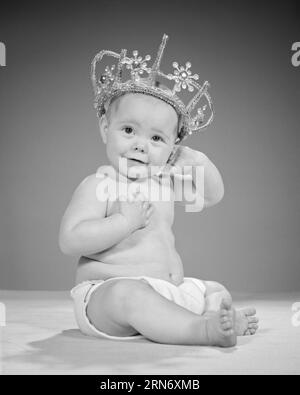 1960ER JAHRE BABY MÄDCHEN SITZEND, DIE KAMERA ANSCHAUEND MIT EINER DIADEM KRONE PRINZESSIN - B21628 HAR001 HARS S S&W AUGENKONTAKT BIZARR HUMORVOLLES GLÜCK SELTSAME GROTESKE, KOMISCHE, UNKONVENTIONELLE WINDELN, DIE EIN LÄCHELN AUF DEN KOPF STELLEN, KONZEPTIONELLE COMEDY-KOPFBEDECKUNG, FRÖHLICH, ABGEDREHTE, EIGENWILLIGE, JUWELENBESETZTE KÖNIGSHÄUSER, DIE SICH AMÜSANT UND EXZENTRISCH FÜHLEN GROWTH JUVENILES POSIERT REGAL BABY MÄDCHEN SCHWARZ-WEISS KAUKASISCHE ETHNISCHE ZUGEHÖRIGKEIT ERRATISCHE HAR001 ALTMODISCHE UNVERSCHÄMTE PRINZESSIN Stockfoto