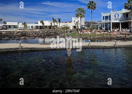 Statue der menschlichen Figur als Wasserbrunnen in einem kleinen See, mit Fischen im Vordergrund, Bars und Hotels im Hintergrund. Marina Rubicon, Lanzarote Stockfoto